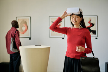 Portrait of young woman engaging with virtual reality headset in modern art gallery with abstract paintings and another visitor in the background