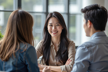 young Asian woman seated in a bright, modern office smiling at two people, a man and a woman, who are seated across from her.
