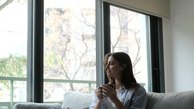 A young woman seated comfortably on her sofa, holding a warm cup of coffee, gazing peacefully out the window at the urban cityscape, enjoying her private moment of relaxation and planning