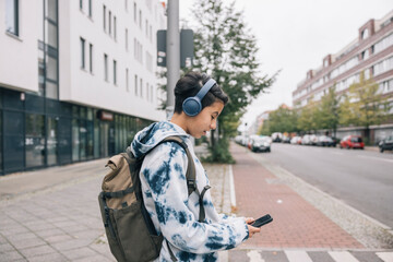Teenager wearing a tie-dye hoodie, backpack, and headphones, looking at a smartphone on an urban...
