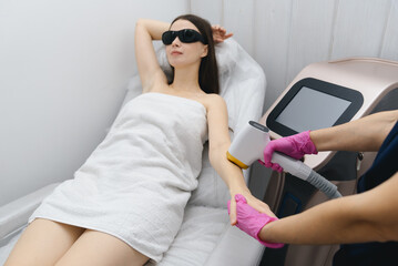 laser epilation, hair removal procedure, young beautiful caucasian smiling brunette woman lying on couch in beauty salon, arm epilation, pink gloves, black glasses, spa and cosmetology concept
