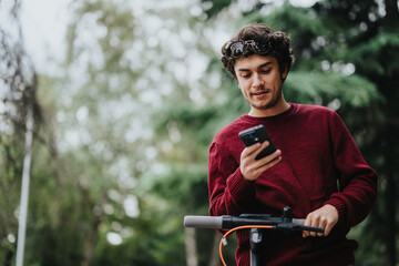 A young man in a red sweater uses his smart phone while standing with an electric scooter in a park...