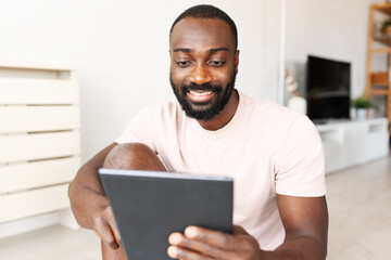 Young African-American man sitting on a yoga mat at home after exercising, smiling while using a tablet for a wellness app in a bright, plant-filled space