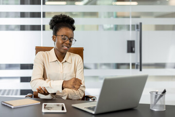 Businesswoman feels arm pain while working at a laptop in modern office. Woman in glasses, formal attire supports elbow joint, indicating discomfort. Contemporary workspace fosters productivity