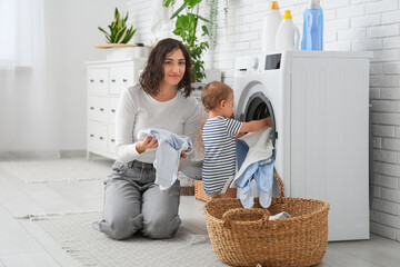 Young mother and her cute little baby putting clothes into washing machine in laundry room