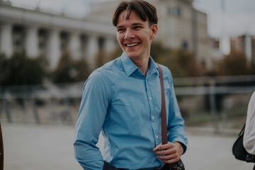 A cheerful young professional man in a blue shirt smiles as he stands outdoors in an urban setting, radiating confidence and positivity.