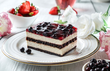 Piece of delicious mousse cake with berry jelly on plate over grey background with cup of juice, berries and flowers. Holiday sweets, dessert and pastry, close up