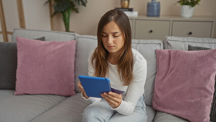 Young attractive woman holding a credit card while using a tablet, sitting on a couch in a cozy living room.