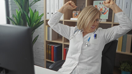 Obraz premium A smiling middle-aged blonde woman in a clinic office, wearing a white medical coat and stethoscope, raises her arms in excitement.