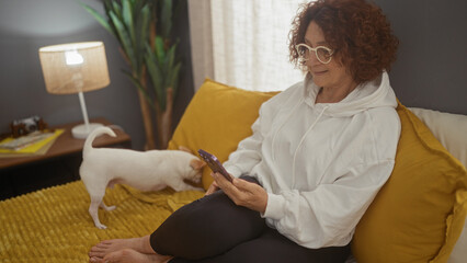 An elderly woman with glasses sits on a bed with a yellow blanket in a bedroom, using her smartphone while a chihuahua dog plays nearby.