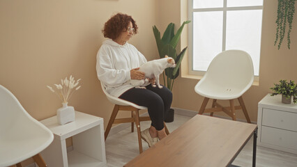 An elderly woman in a hoodie sits in a modern, minimalistic indoor living room holding a small chihuahua dog on her lap.