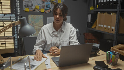 A focused young woman detective works in a cluttered office, analyzing documents beside her laptop.