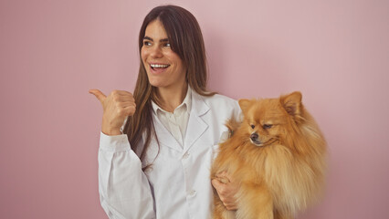 Smiling young woman holding a pomeranian dog points to the side, isolated on a pink background.