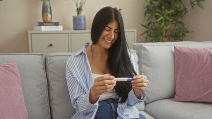 Young hispanic woman with long dark hair smiling while looking at a pregnancy test in the living...