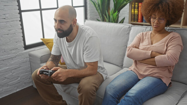 An interracial couple in a living room, man playing video games and woman upset, depicting relationship issues or leisure time conflicts.