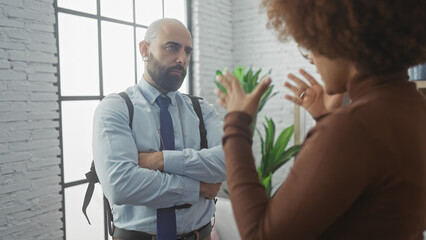 A bald man and an african american woman engaged in a serious conversation indoors with a modern window backdrop.