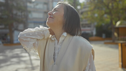 Middle-aged woman with short hair experiencing neck pain in an urban park setting.