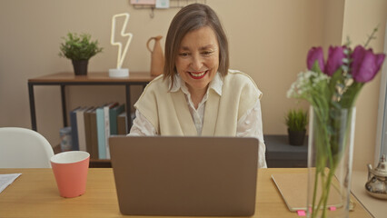 Smiling middle-aged woman using laptop at home office with flowers and modern decor