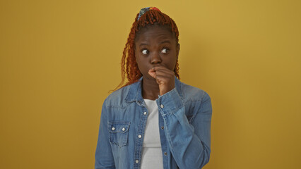 A thoughtful young african american woman in a denim jacket poses against a solid yellow background, conveying contemplation.