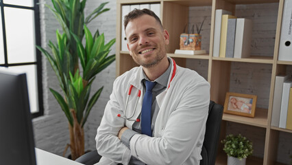 A smiling confident man with a beard wearing a white lab coat and stethoscope sitting in a modern clinic office.