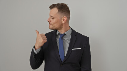 Handsome young man with beard in business suit pointing with thumb against isolated white background
