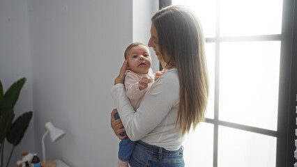 Woman holding baby lovingly in bright living room with sunlight streaming through window, conveying warmth and family bond in a modern indoor setting.