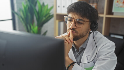 A pensive arab man with a beard and glasses wearing a headset and lab coat in a modern office...