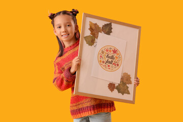 Cute little happy Asian girl holding frame with autumn painting on yellow background