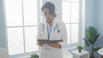 Mature hispanic woman in a white lab coat uses a tablet in a bright hospital room.