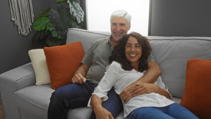 Obraz premium Middle-aged couple smiling together on a cozy sofa in a stylish living room with plants and soft cushions, showcasing a loving relationship in a comfortable home setting.