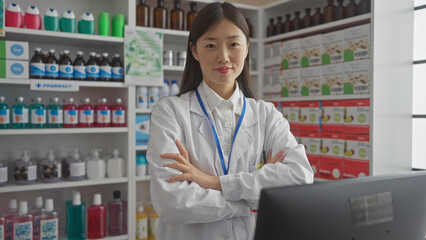 A confident asian woman pharmacist with crossed arms standing in a pharmacy full of medicine shelves.