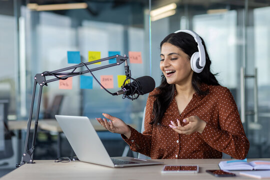 Latin American woman businesswoman recording podcast with microphone and laptop, wearing headphones. She's in a modern workspace, expressing enthusiasm. Smartphone visible on desk.