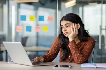 Latin American businesswoman appears stressed at work, gazing at laptop screen in modern office. Creative ideas on sticky notes surround her, with smartphone by her side, reflecting focus.