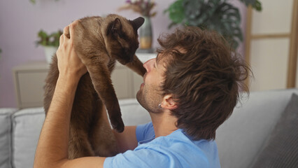 A young hispanic man lovingly interacts with his siamese cat at home.