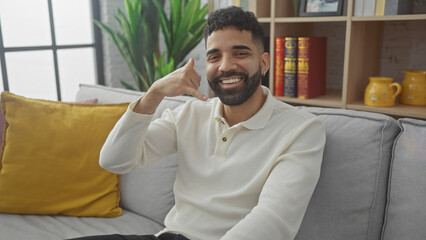 Smiling hispanic man with beard sitting on a couch in a modern living room, making a call me gesture