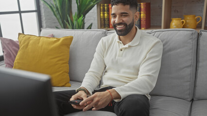 A cheerful young hispanic man with a beard comfortably seated on a gray couch in a cozy living room, holding a remote control.