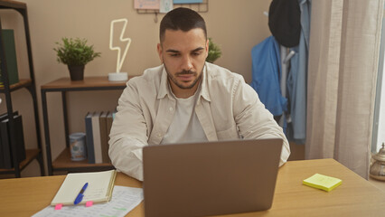 A focused hispanic man works on a laptop at home, showcasing a modern living space and casual attire.