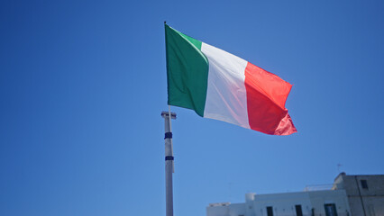 The italian flag waves under a clear blue sky in the coastal town of monopoli, italy, capturing the essence of european outdoor scenery.