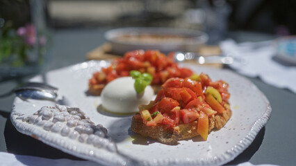 A close-up of fresh bruschetta with tomatoes, basil, and mozzarella served on a white plate outdoors with natural light.