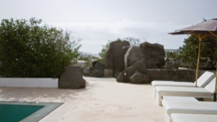 Blurred tropical resort showing out-of-focus lounge chairs by a pool, surrounded by lush greenery and rocks under a clear sky.
