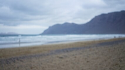 Blurred image of a beach with defocused people, a hazy coastline, and soft waves crashing under an overcast sky creating a serene, dreamy atmosphere