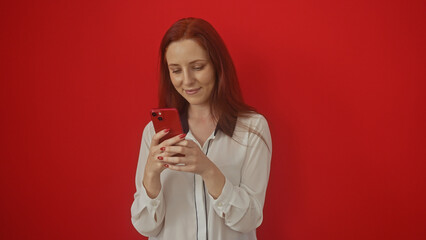 A smiling young woman with red hair using a smartphone against a red background.