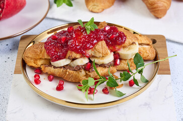 Croissant with banana pieces, chocolate paste and pomegranate seeds in a round white plate