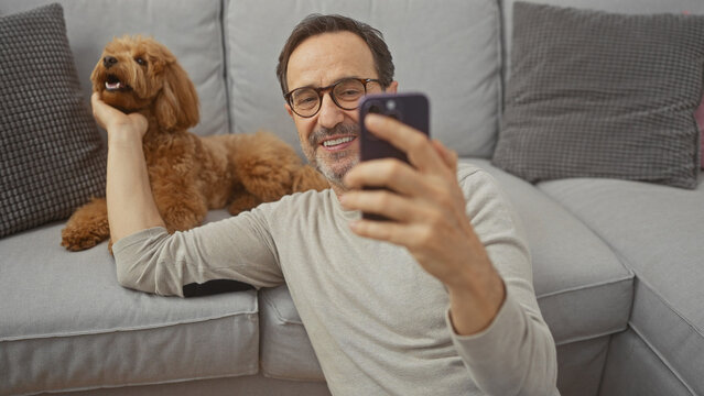 A smiling middle-aged man takes a selfie with a poodle in a cozy living room setting.