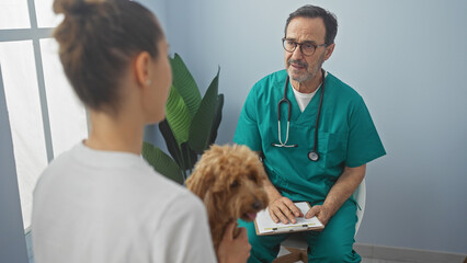 A veterinarian in scrubs consults with a woman holding a poodle in a clinic room interior, implying healthcare for pets.