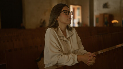 A young hispanic woman is seen in a contemplative pose within a dimly lit christian church in...