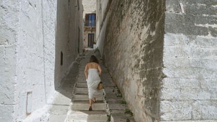 A beautiful young woman in a white dress climbs narrow steps in the old town of ostuni, puglia, italy, europe, capturing the essence of mediterranean charm and historic architecture.