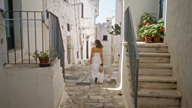 A young, beautiful hispanic woman walks through the charming old town of ostuni in puglia, italy, surrounded by whitewashed buildings and cobblestone streets.