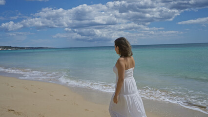 A young, beautiful hispanic woman in a white dress enjoys a serene walk along the sandy beach of pescoluse, salento, puglia, italy, with the clear blue sea and cloudy sky in view.