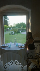 A beautiful young hispanic woman enjoys an indoor dining experience at a charming restaurant in italy, seated at a table elegantly set for two with a scenic garden view.